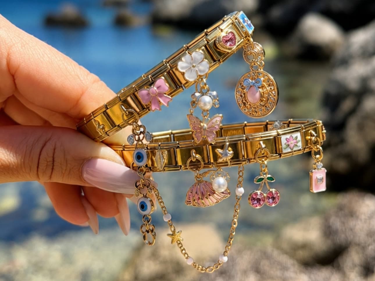 Zwei goldene italienische Charm-Armbänder am Wasser in die Höhe gehalten, mit Schmetterling, Kirschen, Schleife, Muschel und Evil Eye